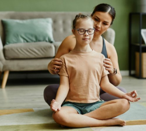 Portrait of female instructor assisting young girl with down syndrome doing yoga and meditating in fitness studio, copy space